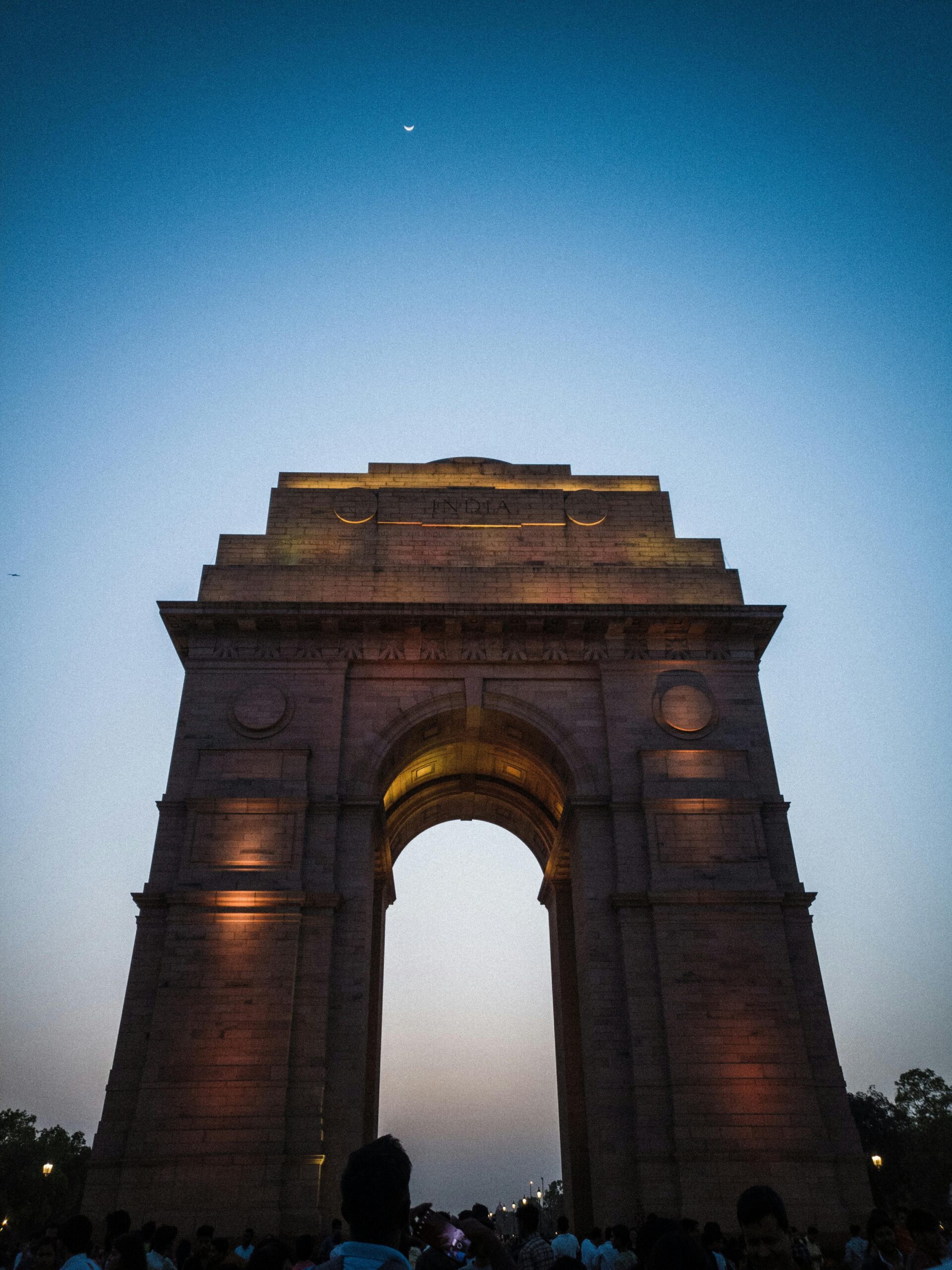 Nighttime view of the illuminated India Gate in New Delhi against a clear sky with crescent moon.
